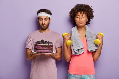 Hombre con un pastel en las manos y mujer practicando deporte con unas pesas evitando comer el pastel